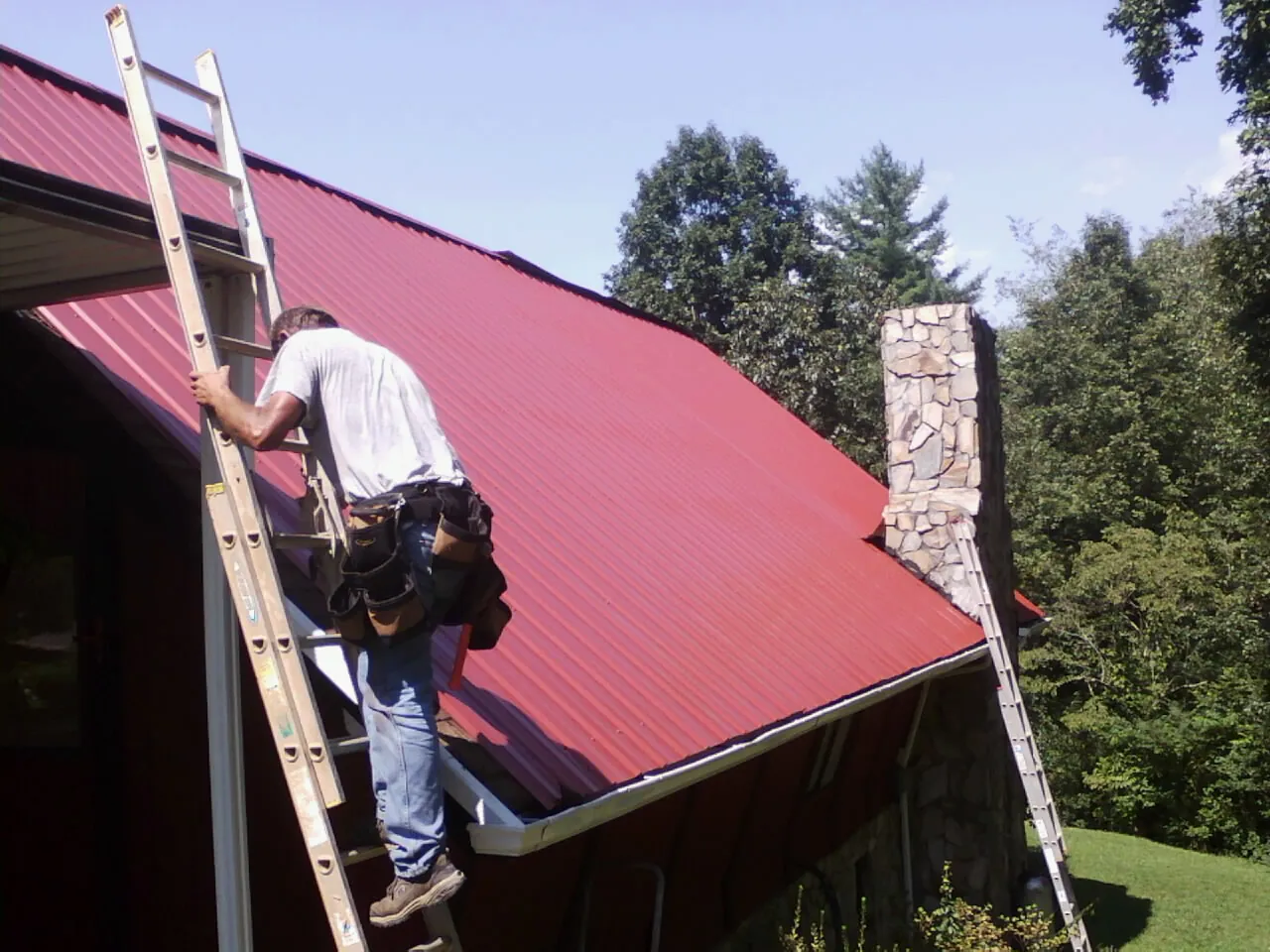 Roofer on a ladder beside a red metal roof and stone chimney — Soffit & Fascia Repair by Roofing Brothers in Sneads Ferry